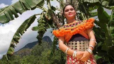 seorang gadis orang asli Sarawak dalam pakaian tradisi budaya berdiri di hadapan pokok pisang. Di latar belakang adalah bukit dan pokok.