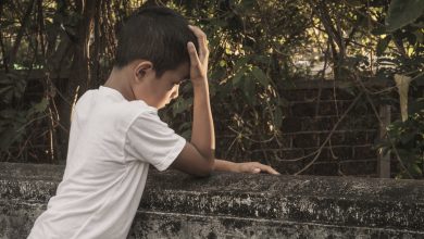 A boy stands against the wall with his head in his palm. The boy looks sad.