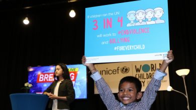 A boy holds up a placard that says If children ran the UN, 3 in 4 will work to end violence and bullying for every child. Behind him a girl presenter stands infront of a TV screen with the message Breaking News.