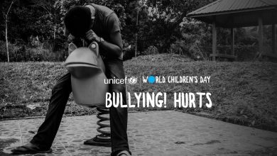 A dejected looking boy sits in the playground with his head on his hand. On the photo is the phrase Bullying hurts!