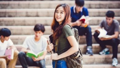 Uncovering school hacks with a college student holds her books. Behind her are some other students sitting on the stairs.