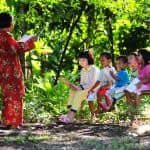 A teacher teaches four children sitting on a bench in the outdoors.