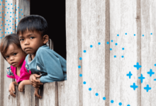 Two migrant children in Malaysia - a girl and a boy - look out of window of a wooden house.